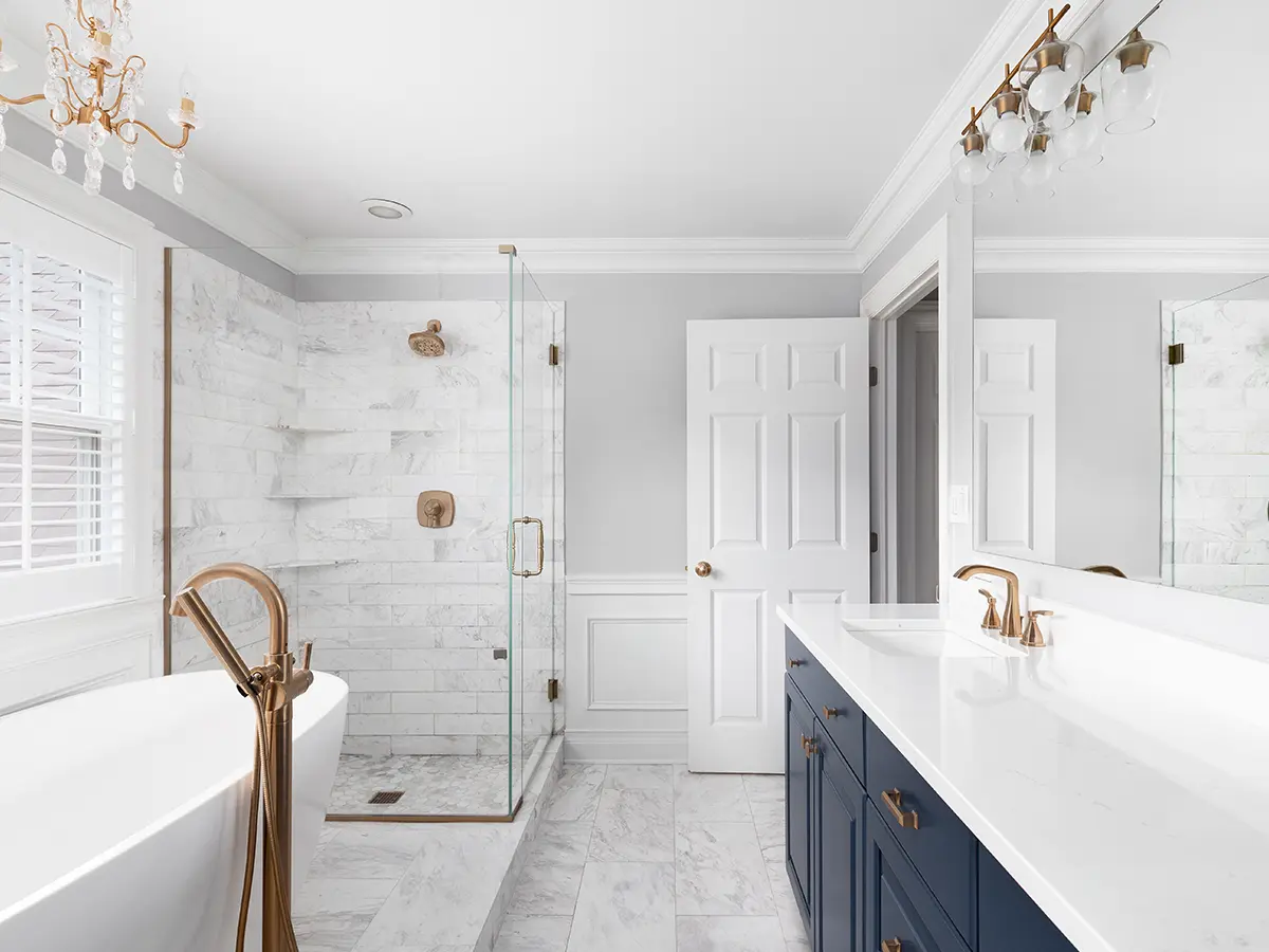 An upscale bathroom with a navy blue double vanity, glass walk-in shower, and freestanding tub with golden water fixtures