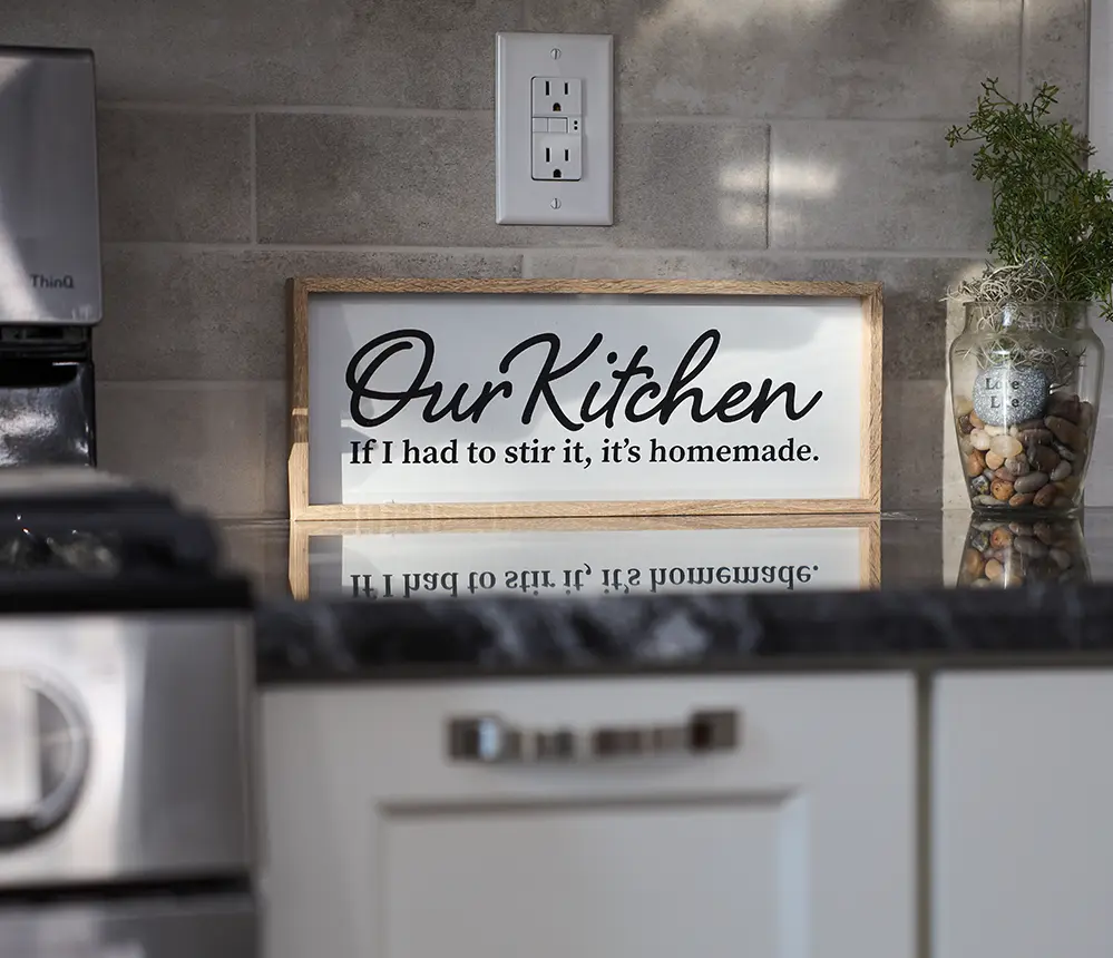A kitchen with a painting that reads "our kitchen, if i had to stir it, it's homemade."