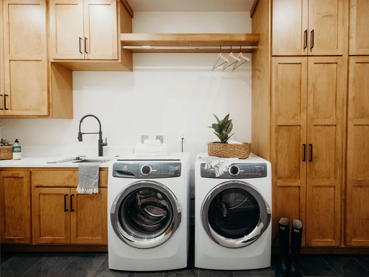 Modern laundry room with front-load washer and dryer, light wood cabinetry, and black faucet.