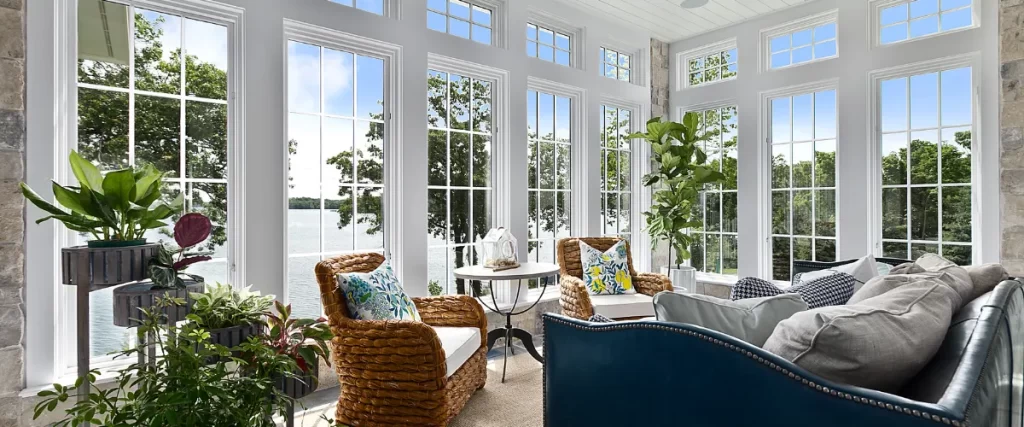 A bright sunroom with floor-to-ceiling windows overlooking a lake, decorated with wicker furniture, plush cushions, and lush indoor plants.